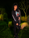 Full length shot of female model with black hair standing in a spooky cemetery and wearing the BEETLEJUICE™ Lydia Strange Long Sleeve Tee. 