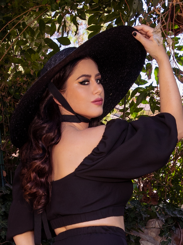 Ashley facing away from the camera, but looking back at it over her shoulder, wears the Lydia Sun Hat from gothic glamour clothing brand La Femme en Noir.