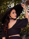 Ashley facing away from the camera, but looking back at it over her shoulder, wears the Lydia Sun Hat from gothic glamour clothing brand La Femme en Noir.
