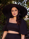 Ashley, shot from the waist up, wears an all black outfit highlighted by the Lydia Sun Hat.