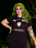 Green-haired model wearing the BEETLEJUICE™ Lydia Dead Tee while posinng in a graveyard.