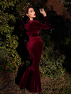 A back shot of Micheline Pitt in a lush garden looking into the sunlight while modeling the Black Marilyn gown in oxblood.