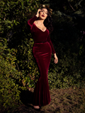 A full length photo of Micheline Pitt in a lush garden looking into the sunlight while modeling the Black Marilyn gown in oxblood.