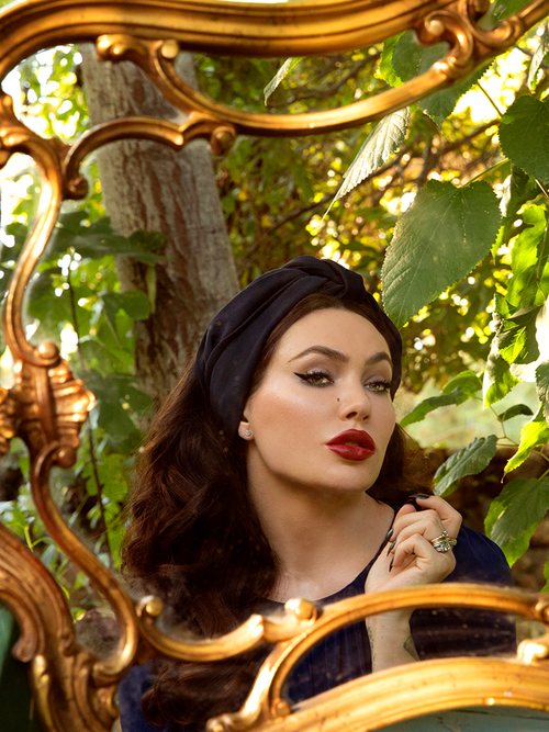 Micheline Pitt gazing into a gold plated mirror wearing the Art Deco Head Scarf in Navy Ponte.