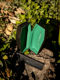The Vamp Coffin Sunglass Case in Emerald Green pictured sitting on a tree stump among a sea of moss.