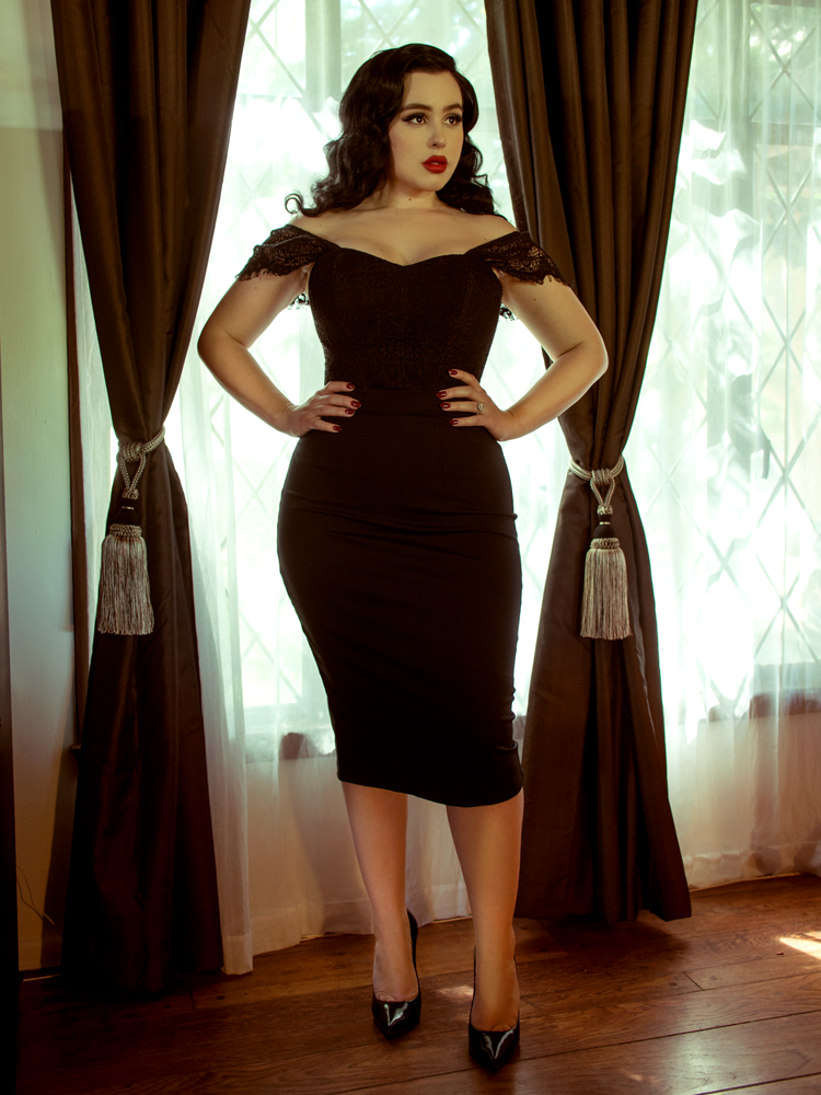 Rachel standing in front of a window with her hands on her waist modeling the Vamp pencil skirt in black by La Femme En Noir.