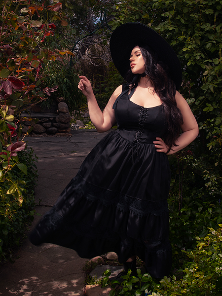Bathed in sunlight within a lively garden, a brunette beauty graces the scene, captivating onlookers with her style in the striking Pickety Witch Dress in Black, a gothic dress creation by La Femme en Noir.