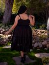 Within the enchanting garden's embrace, a radiant brunette muse becomes a captivating sight, donning the exquisite Pickety Witch Dress in Black from La Femme en Noir, a stunning gothic dress that demands attention.