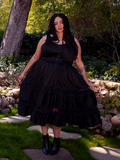 Bathed in sunlight within a lush garden, a brunette beauty enchants with her presence, adorned in the mesmerizing Pickety Witch Dress in Black, a remarkable gothic dress crafted by La Femme en Noir.