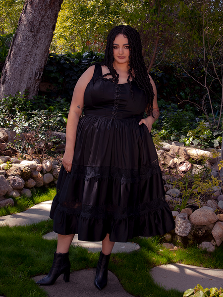In the embrace of a sunlit garden, a mesmerizing brunette figure exudes enchantment, clad in the captivating Pickety Witch Dress in Black from La Femme en Noir, a gothic dress that mesmerizes all.