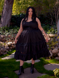 Under the golden rays of the sun, a brunette model flourishes in a vibrant garden, showcasing the striking Pickety Witch Dress in Black, a remarkable gothic dress creation by La Femme en Noir.