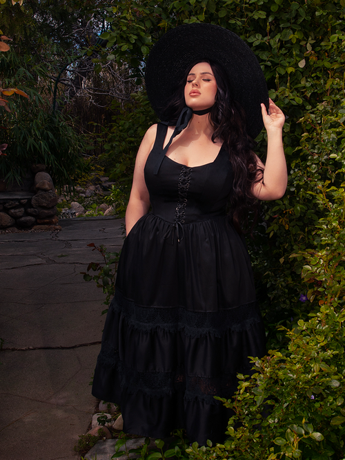Under the radiant sun in a vibrant garden, a brunette model radiates beauty while donning the mesmerizing Pickety Witch Dress in Black, a stunning gothic dress by La Femme en Noir.