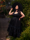 In the midst of a sun-drenched garden, a brunette muse shines brightly, showcasing the allure of the Pickety Witch Dress in Black from La Femme en Noir, a perfect embodiment of gothic fashion.