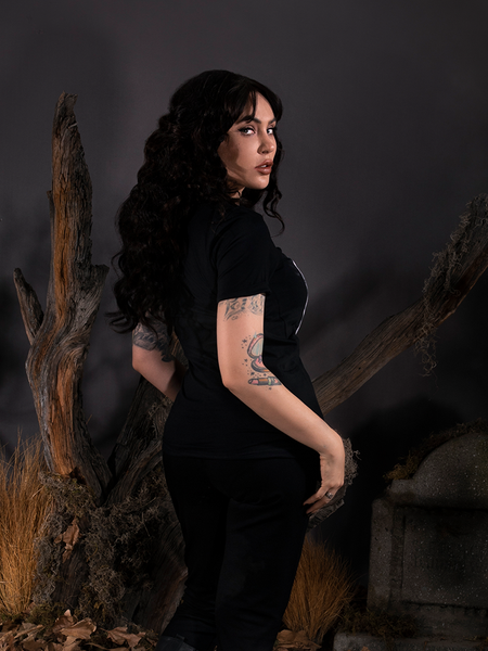Posing in a spooky cemetery scene, Micheline Pitt turns away from the camera but looks back at it while wearing the Sleepy Hollow™ Heads Will Roll Tee.