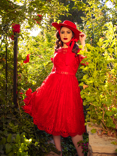 Female model stands in a red rose garden while wearing the the Mourning Dress in Crimson Lace from goth clothing brand La Femme en Noir.