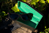 The Serpent Sunglasses in Black sitting on top of the opened coffin-shaped carrying case along with branded cleaning cloth.