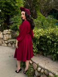 Turned away from the camera, Micheline Pitt looks back while in the Oxblood Salem Dress from gothic dress retailer La Femme en Noir.