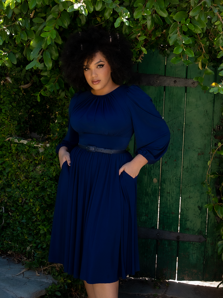 Model Ashleeta poses in the Salem Dress in Navy while standing in front of a green door covered in lush greenery.