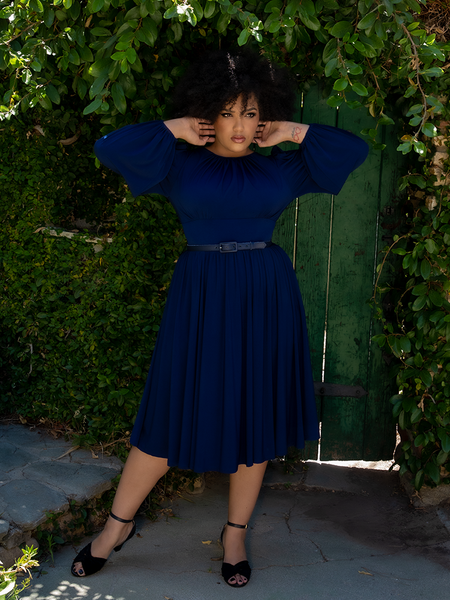 Ashleeta poses in front of a green garden door while wearing the Salem Dress in Navy. 