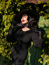 Micheline Pitt, with the glow of the sun on her face, pulls on the bow of her black witch hat while modeling the Salem Top in Black.