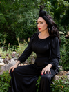 Micheline Pitt sitting on a stone bench covered in green moss while modeling the Salem Top in Black.