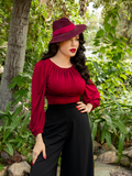 Micheline Pitt standing in her garden while wearing a oxblood retro style gothic top.