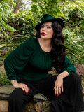 Micheline Pitt sat in a lush, green garden with black flowing pants, and matching hunter green top and hat.