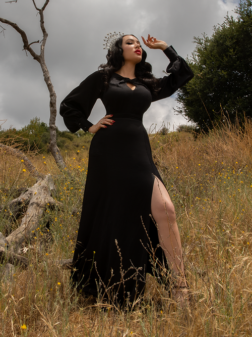 Standing against a grey skies in a field with dead trees, Micheline Pitt models the goth style Opera Satin Gown in Black.