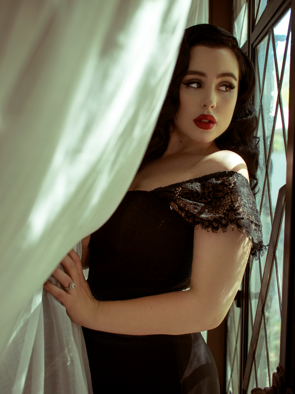 Rachel leans on a window and pulls back a white curtain while modeling the Southern Gothic bustier top in black.