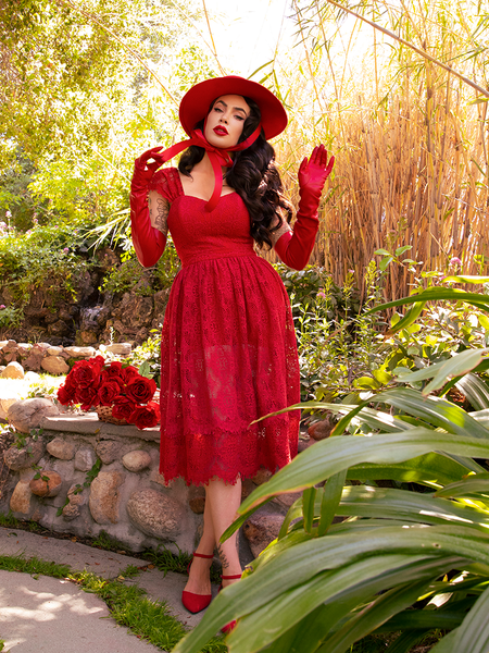 Micheline Pitt standing in a lush garden while wearing an all crimson gothic style outfit.