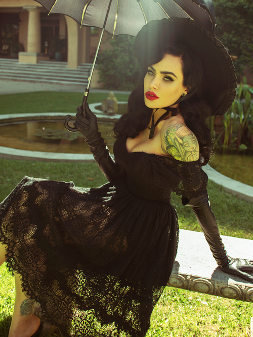 Micheilne Pitt sitting on a stone bench holding a black parasol models a gothic inspired outfit from La Femme en Noir.