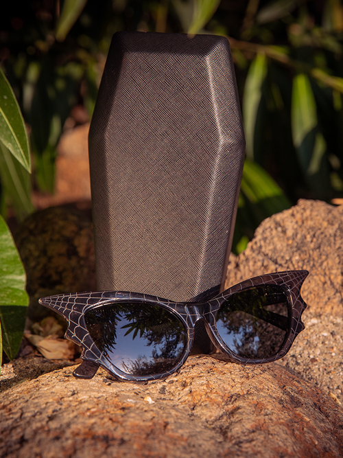Isolated product shot of the Vamp Batwing Sunglasses in Smoke Spider Web from Vixen Clothing sitting in front of their coffin carry case.