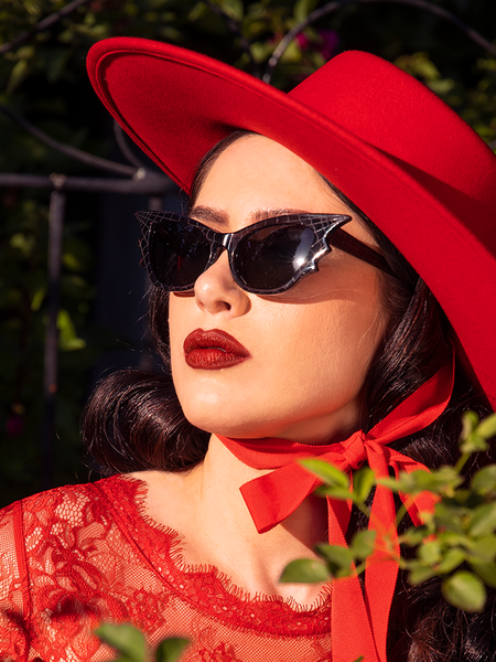 Ashley stares into the sun while wearing the Vamp Batwing Sunglasses in Smoke Spider Web with an all red goth vintage outfit.