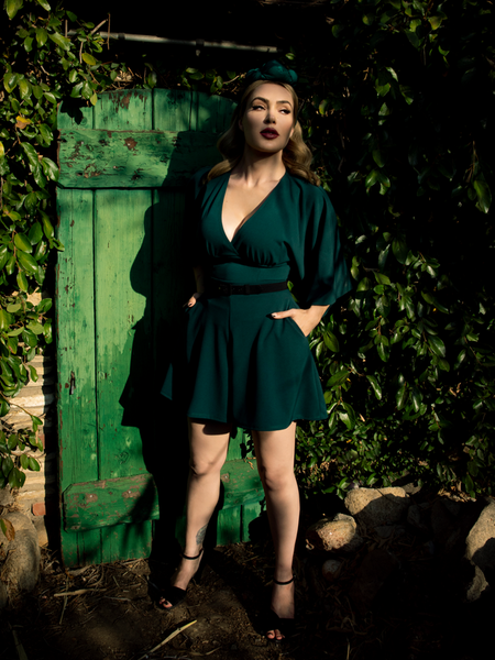 A full length shot of Micheline Pitt, with her hands in her pockets standing in a garden, models the Black Widow tap shorts in hunter green from La Femme En Noir.