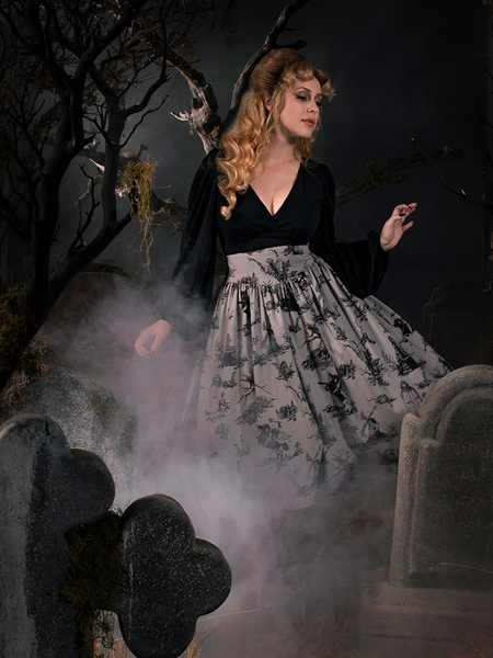 Linda swaying in a graveyard while wearing a gothic vintage outfit including a flowy black longsleeve top and Sleepy Hollow Gothic Tale Toiles Skirt in Grey.