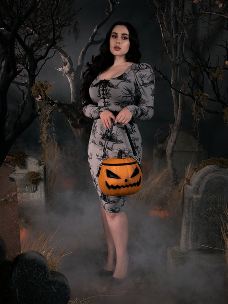 Rachel Sedory standing in a haunted graveyard in a gothic dress while holding a pumpkin bag.