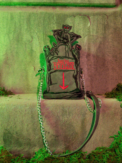 The BEETLEJUICE™ Tombstone Crossbody Bag photographed on a mossy step.