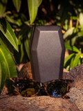 Product shot of the Vamp Batwing Sunglasses in Tortoise positioned in front of the included coffin carrying/storage case.
