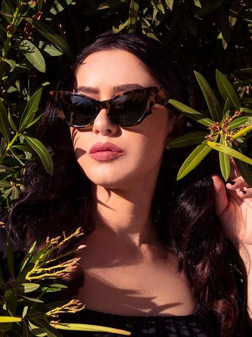 Ashley standing amongst a lush, green background wears the Vamp Batwing Sunglasses in Tortoise.