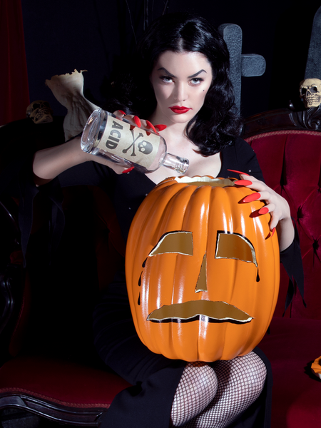 Healther Black pouring from a bottle labeled "Acid" into a carved pumpkin while wearing the Vampira® Show Gown.
