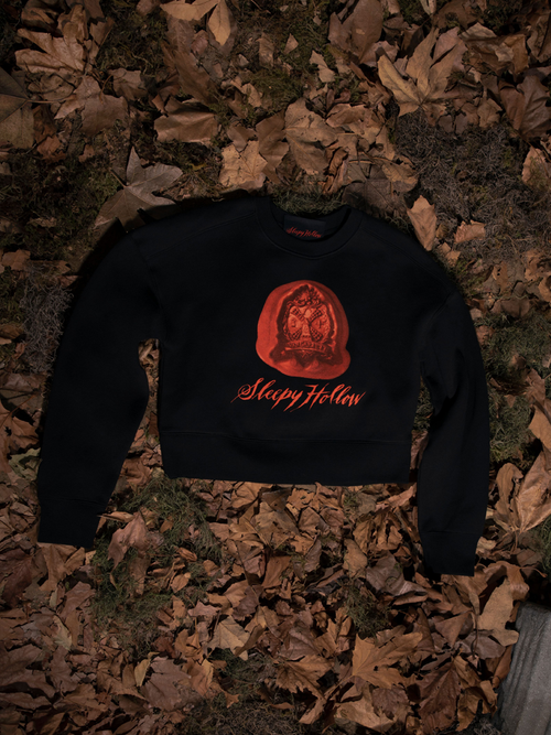 The Sleepy Hollow™ Van Garrett Wax Seal Cropped Sweatshirt laid over a bed of dead leaves.