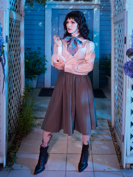 Stephanie adjusting the sleeve on her long-sleeve white shirt while standing in a spookily decorated scene in front of a Victorian house while showcasing the Tim Burton's CORPSE BRIDE™ Victor Pinstripe Skirt in Dusty Olive from goth glamour clothing company La Femme en Noir.