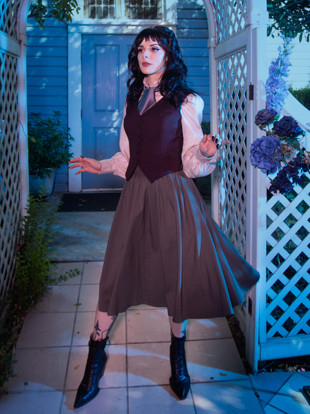 Stephanie poses with her arms slightly outstretched while standing under a white archway adorned with blue and purple flowers while modeling the Tim Burton's CORPSE BRIDE™ Victor Pinstripe Skirt in Dusty Olive from gothic retro clothing company La Femme en Noir.