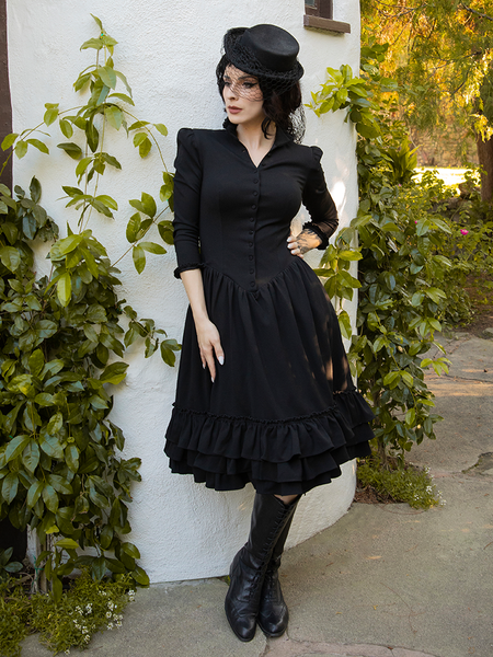 Goth model poses with one hand on her hip and the other resting at her side while modeling the Victorian Dress in Black from goth dress brand La Femme en Noir.
