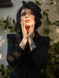 A raven haired model holds her hands up on either side of her face while modeling the Victorian Dress in Black from goth clothing brand La Femme en Noir.