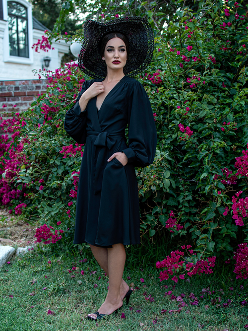 A full length shot of Aliza, standing in a garden, wears the Serpentine wrap dress in black from La Femme En Noir.