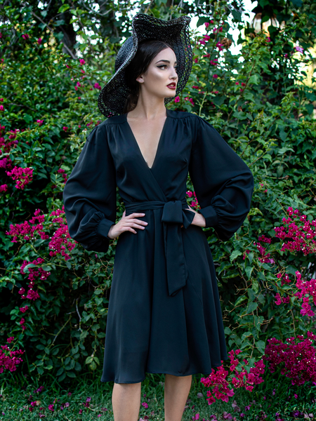 Aliza, standing in a garden, wears the Serpentine wrap dress in black from La Femme En Noir.