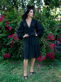 Aliza, with her hands on her hips standing in a garden, wears the Serpentine wrap dress in black from La Femme En Noir.