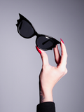 The Vamp Batwing Sunglasses being held up by a fair skinned hand with long red nails.