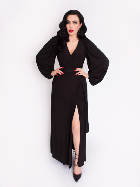 Micheline Pitt standing in front of a white backdrop modeling the all black Black Widow Wrap Gown in Solid Black